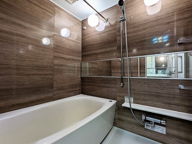 木目調の鏡面パネルの浴室です。照明が壁にうつり高級感があります。鏡は横に長いです。浴槽は白で床も白です。