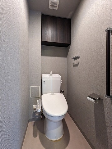 床は淡いブラウン、壁はグレーのクロスです。温水洗浄機能付きトイレでトイレ上部にダークブラウン縦木目調の扉付き収納があります。
